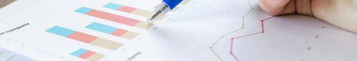 A close-up of a hand with a pen analyzing data on colorful bar and line charts on paper.